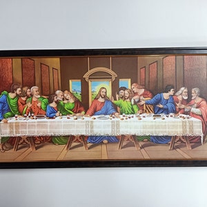 Op de afbeelding: Een ingelijst schilderij dat het Laatste Avondmaal voorstelt, een beroemde bijbelse scène. Het schilderij toont Jezus Christus en zijn twaalf discipelen die rond een tafel zijn verzameld, met een gedetailleerde achtergrond en levendige kleuren.