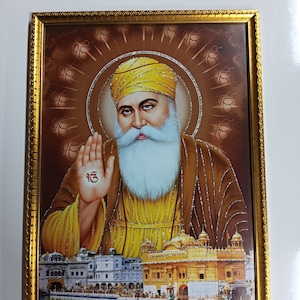 May include: A framed religious print featuring a portrait of a man with a long white beard and a yellow turban. The man is holding his hand up in a gesture of blessing. The background of the print is brown with a gold border. The print also features a golden temple in the background.