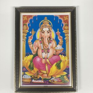 May include: A framed image of the Hindu deity Ganesha, depicted in a colorful, traditional style. He is seated on a lotus flower, holding a sweet in his right hand and a modaka in his left. The image is surrounded by a brown frame with gold accents.
