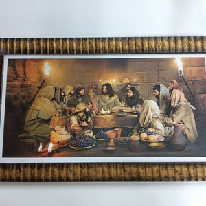 Op de afbeelding: Een ingelijst schilderij dat het Laatste Avondmaal voorstelt, een bijbelse scène waarin Jezus een maaltijd deelt met zijn discipelen. Het schilderij speelt zich af in een zwak verlichte kamer met stenen muren en toont een tafel die beladen is met eten en wijn. De figuren zijn gekleed in traditionele gewaden en zijn verzameld rond de tafel, met Jezus in het midden.