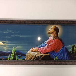 Puede incluir: Una obra de arte religiosa enmarcada que representa una figura con túnicas rojas y azules, mirando hacia arriba con un halo. El fondo muestra una escena nocturna con una luna, nubes y edificios, en un marco marrón oscuro.