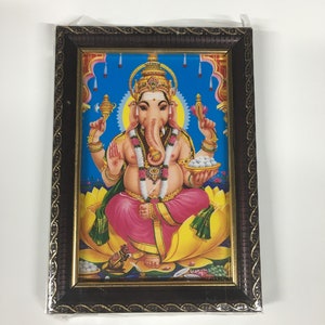 May include: A framed picture of a Hindu deity, Ganesha, with a blue background. Ganesha is depicted with an elephant head, four arms, and a pink and yellow outfit. He is sitting on a yellow lotus flower. The frame is brown with gold accents.