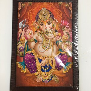 May include: A framed painting of the Hindu deity Ganesha, depicted with an elephant head, four arms, and a large belly. He is seated on a throne and holding a variety of objects, including an axe, a lotus flower, and a sweet. The painting is set against a background of gold and red.