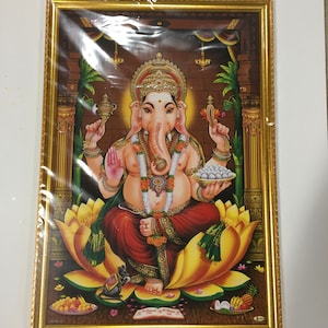May include: A framed print of a Hindu deity, Ganesha, depicted in a colorful, ornate style. He is seated on a lotus flower, holding a sweet in his hand and a bowl of sweets in his other hand. A rat sits at his feet.