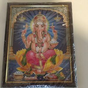 May include: A framed print of a Hindu deity, Ganesha, depicted in a colorful, ornate style. He is seated on a lotus flower and holding a variety of offerings, including sweets and fruits. The background is a blue and gold design with a halo of light around Ganesha's head.
