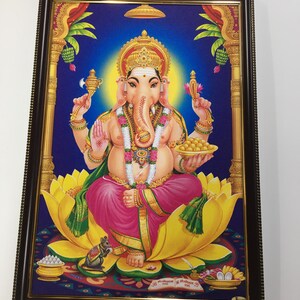 May include: A framed print of a Hindu deity, Ganesha, depicted with four arms, a trunk, and a large belly. He is seated on a yellow lotus flower and holding a variety of objects, including a sweet treat, a mirror, and a pot. The background is blue with a golden border.