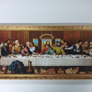Op de afbeelding: Een ingelijste prent van "Het Laatste Avondmaal" met Jezus en zijn discipelen aan een tafel. Het kunstwerk heeft een gouden, sierlijke lijst, een wit tafelkleed en figuren in gewaden. De achtergrond is warmbruin met architecturale details.