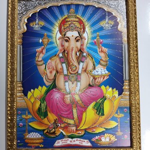 May include: A framed image of the Hindu deity Ganesha, depicted with a blue background, sitting on a lotus flower. He is holding a sweet in his right hand and a mirror in his left hand. The image is surrounded by a gold frame with intricate details.