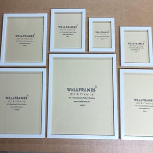 May include: A collection of seven white picture frames of varying sizes, arranged on a neutral surface. Each frame has a white border and a light-colored backing. The frames are labeled with the text "WALLFRAMES Art & Framing".