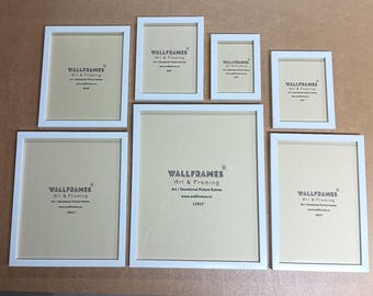 May include: A collection of seven white picture frames of varying sizes, arranged on a neutral surface. Each frame has a white border and a light-colored backing. The frames are labeled with the text "WALLFRAMES Art & Framing".