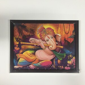 May include: A framed painting of a Hindu deity, Ganesha, playing a flute. The painting is set against a colorful background with flowers and foliage. Ganesha is depicted with a red and gold turban, a pink body, and a large trunk.