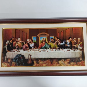 Op de afbeelding: Een ingelijst schilderij dat het Laatste Avondmaal voorstelt, een beroemde Bijbelse scène. Het schilderij toont Jezus Christus en zijn twaalf discipelen die rond een tafel zijn verzameld, met een gedetailleerde weergave van de kamer en de tafeldekking.
