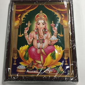 May include: A framed print of a Hindu deity, Ganesha, depicted in a colorful, ornate setting. He is seated on a yellow lotus flower, holding a sweet in his hand, and surrounded by offerings. The frame is brown with gold accents.