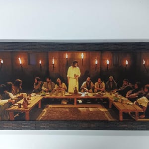 Op de afbeelding: Een ingelijste prent die het Laatste Avondmaal uitbeeldt. De afbeelding toont een centrale figuur in een gele jurk die voor een lange houten tafel staat met anderen eromheen. Fakkels verlichten de scène tegen een donkere achtergrond.