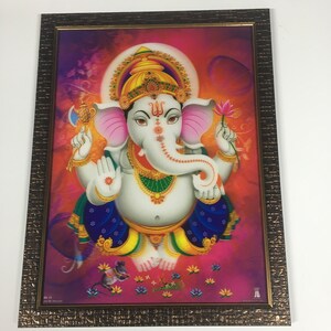 May include: A framed print of a Hindu deity, Ganesha, with a pink background. Ganesha is depicted with a white body, a pink trunk, and four arms. He is holding a lotus flower in one hand and an axe in another. He is wearing a gold crown and a blue and yellow robe. The frame is brown with a gold trim.