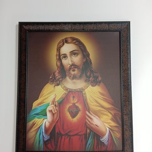 May include: A framed painting of Jesus Christ with a golden halo, wearing a red robe with a yellow cloak, and his hands clasped over his heart. The heart is depicted as a red, flaming heart with a crown of thorns and a cross.