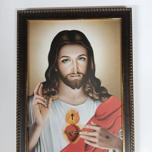 Peut inclure: Une estampe religieuse encadrée représentant Jésus-Christ portant une robe rouge et un cœur enflammé avec une couronne d'épines. L'image est sur un fond clair avec des rayons de lumière émanant de derrière Jésus.