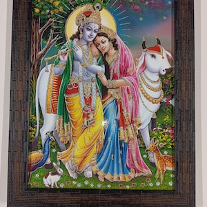 May include: A framed artwork depicting a couple in traditional Indian attire, surrounded by a white cow, peacock, rabbit, and deer. The couple is adorned with jewelry and a halo. The artwork is set against a backdrop of a lush landscape.