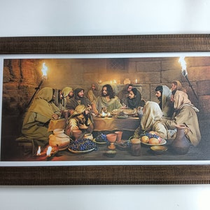 Op de afbeelding: Een ingelijste prent die het Laatste Avondmaal uitbeeldt. Het kunstwerk toont Jezus en zijn discipelen aan een tafel, verlicht door kaarsen en fakkels. De lijst is donkerbruin met een getextureerde uitstraling.