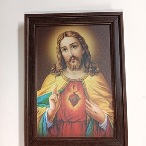 May include: A framed painting of Jesus Christ with a heart on his chest. The heart is red and has a crown of thorns around it. The painting is in a brown frame.
