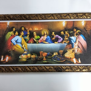 Op de afbeelding: Een ingelijst schilderij dat het Laatste Avondmaal voorstelt, een bijbelse scène met Jezus en zijn discipelen die rond een tafel zitten. Het schilderij speelt zich af in een zwak verlichte kamer met een warme kleurenpalet en toont realistische details.