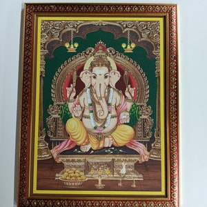 May include: A framed painting of a Hindu deity, Ganesha, with four arms, sitting on a throne with a green background. The painting is surrounded by a gold and red frame.