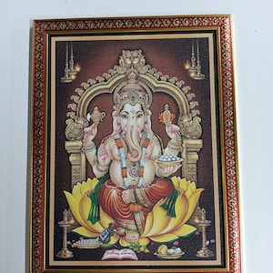 May include: A framed print of a Hindu deity, Ganesha, sitting on a lotus flower. He is depicted with a large head, four arms, and a trunk. He is holding a sweet in one hand and a book in another. The print is surrounded by a gold and red frame.