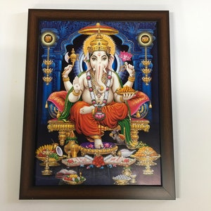 May include: A framed print of a Hindu deity, Ganesha, sitting on a throne with a blue background. He is depicted with a large head, four arms, and a trunk. He is holding a lotus flower, a sweet, and an axe. He is wearing a red and gold robe and a crown. The frame is brown wood.