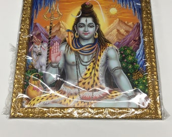 May include: A framed print of a Hindu deity, Shiva, sitting in a meditative pose. He is depicted with a third eye, a snake around his neck, and a bull standing behind him. The print is set against a colorful background of mountains and a sunset.
