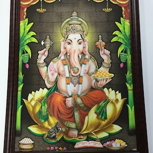 May include: A framed painting of the Hindu deity Ganesha, depicted with an elephant head, four arms, and a large belly. He is seated on a lotus flower and holding a variety of objects, including a sweet treat, a conch shell, and an axe. The painting is set against a dark brown background with a decorative border.