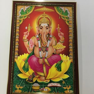 May include: A framed print of a Hindu deity, Ganesha, with an elephant head, sitting on a lotus flower. He is holding a sweet in his right hand and a book in his left hand. The background is red with gold accents.