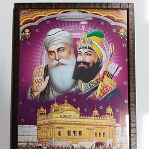 Op de afbeelding: Een ingelijste print van twee Sikh-goeroes, Guru Nanak en Guru Gobind Singh, met een gouden tempel op de achtergrond. De print is op een roze achtergrond met glitteraccenten.