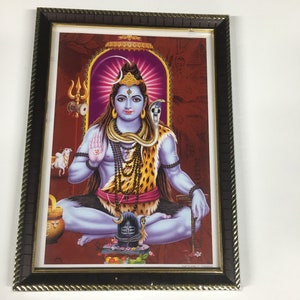 May include: Framed image of a Hindu deity, Shiva, sitting in a meditative pose with a snake around his neck. He is wearing a leopard skin and a beaded necklace. The background is red with gold accents.