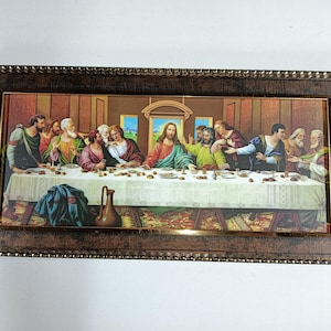 Op de afbeelding: Een ingelijst schilderij dat het Laatste Avondmaal voorstelt, een beroemde bijbelse scène. Het schilderij toont Jezus Christus en zijn discipelen die rond een tafel zijn verzameld, met een gedetailleerde weergave van de kamer en het eten op de tafel.