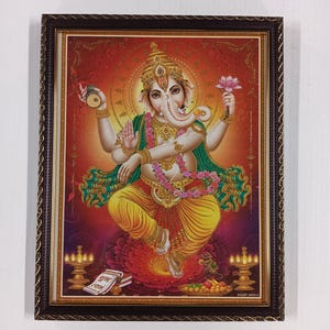 May include: Framed artwork depicting the Hindu deity Ganesha, seated on a lotus flower. Ganesha is shown with four arms, wearing a crown, and holding various symbolic objects. The artwork features vibrant colors and intricate details, with a decorative frame.