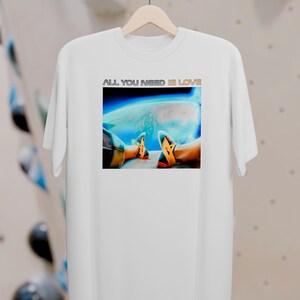 Könnte beinhalten: Weißes T-Shirt mit dem Text "ALL YOU NEED IS LOVE" über einer Grafik von Beinen und Füßen in Turnschuhen, vor dem Hintergrund der Erde aus dem Weltraum. Das T-Shirt hängt an einem Holzbügel.