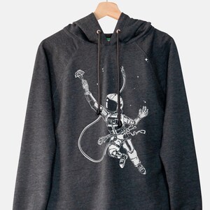 Könnte beinhalten: Dunkelgrauer Kapuzenpullover mit einer weißen Illustration eines Astronauten im Weltraum. Der Astronaut hält einen Fallschirm und ist mit einer Schnur verbunden. Das Design beinhaltet Sterne. Der Hoodie hat eine Kapuze mit Kordelzug und eine Kängurutasche.