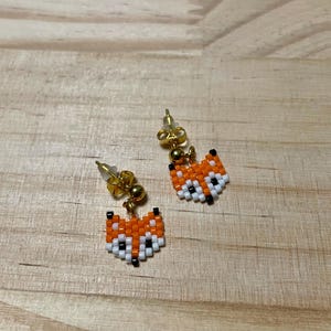 Puede incluir: Un par de pendientes con forma de zorro. Cada pendiente presenta una cabeza de zorro con cuentas en naranja, blanco y negro. Los pendientes tienen postes dorados y pequeñas cuentas doradas. Los pendientes se muestran sobre una superficie de madera.