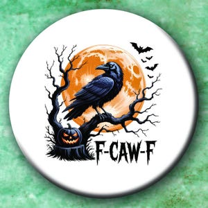 May include: A round, white Halloween-themed button featuring a black crow perched on a bare tree branch in front of a large orange moon. A carved pumpkin and bats are also present. The text "F-CAW-F" is below the tree.