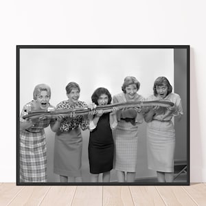 May include: Five women in a black and white photo are holding a long, thin object, possibly a baguette or a loaf of bread, in front of their mouths. They are all smiling and looking at the camera.