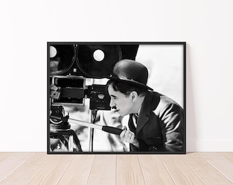 May include: Black and white framed print featuring a vintage photograph of Charlie Chaplin operating a film camera. Chaplin is wearing a bowler hat and suit, focused on the camera. The print is displayed on a light wood floor.