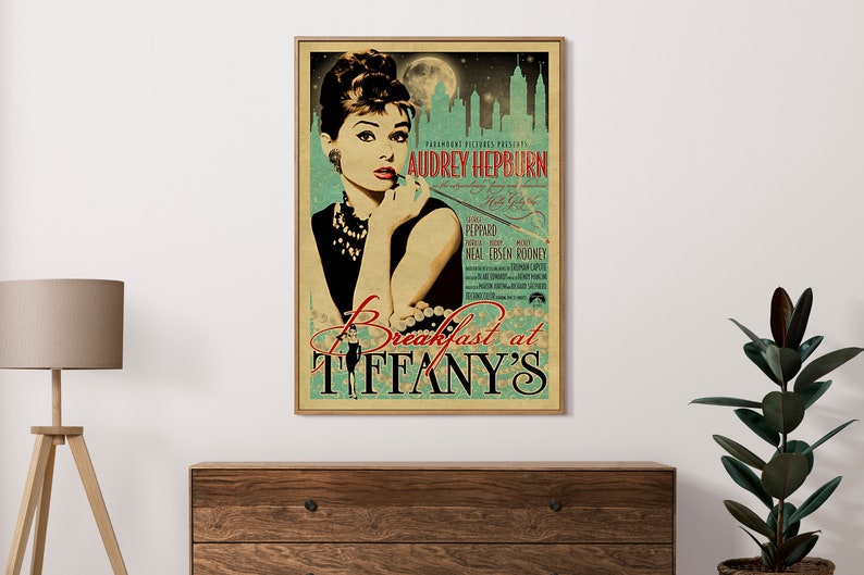 May include: A framed vintage movie poster for "Breakfast at Tiffany's" featuring Audrey Hepburn. The poster has a teal and beige color scheme with the film's title and credits. The image is displayed on a wooden dresser.