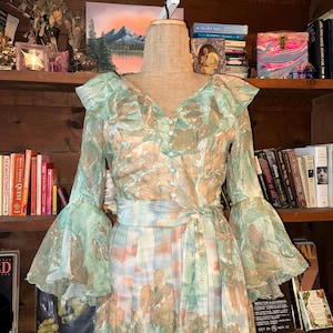 May include: A vintage-style dress with a flowing silhouette, featuring a V-neckline and long, flared sleeves. The dress is adorned with a watercolor-like print in shades of green, peach, and blue. It has a matching belt and is displayed on a mannequin.