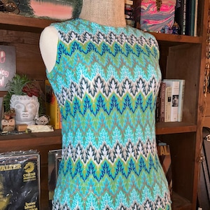 May include: A sleeveless top with a colorful leaf pattern in shades of teal, blue, green, and gold. The vintage garment is displayed on a mannequin, showcasing its intricate design and texture. The top has a round neckline.