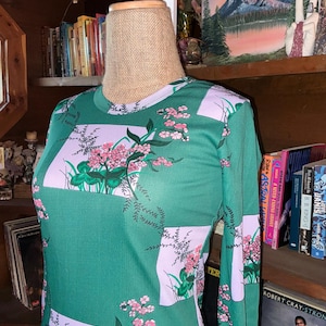 May include: A long-sleeved green dress with a floral pattern. The dress features rectangular panels of light purple with pink flowers and green leaves. The dress is displayed on a mannequin.