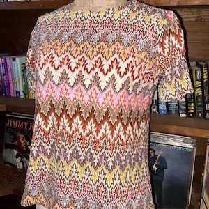 May include: A short-sleeved, patterned top in a variety of colors, including brown, orange, yellow, pink, and purple. The knitwear features a repeating leaf-like design. The top is displayed on a mannequin.