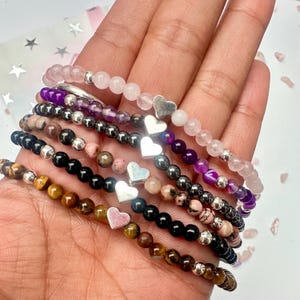 Puede incluir: Una colección de pulseras de cuentas en varios colores, incluyendo rosa, morado, negro y marrón. Cada pulsera presenta pequeñas cuentas redondas y un dije de corazón plateado. Las pulseras se exhiben sobre un fondo claro.