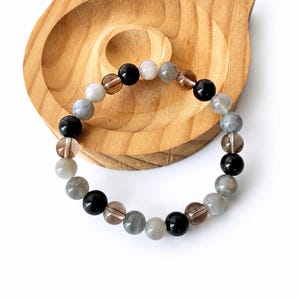 May include: A beaded bracelet featuring a repeating pattern of polished spherical beads in shades of black, translucent brown, and opaque gray. The beads are strung together in a circular arrangement, showcasing a smooth, reflective surface texture.