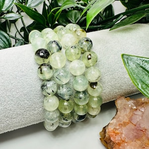 May include: A stack of green and black gemstone bracelets with round beads. The bracelets are made of clear green stones with black inclusions.