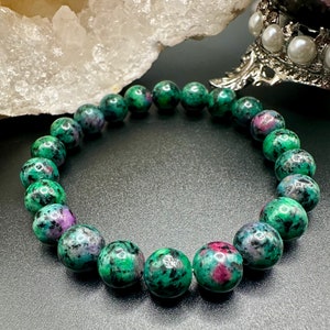 May include: A bracelet made of round gemstones, displaying a mix of green, red, and black hues. The beads are set against a dark backdrop, with a white crystal and a decorative silver stand visible in the background.